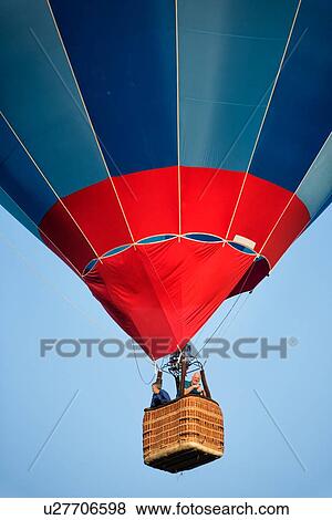Stock Photo - Todi, Italy: July 18, 2013; Todi Mongolfiere Hot Air Balloon Festival - red, blue and navy hot air balloon rising into sky as seen from below, detail view, with passengers in basket, in early morning sunshine, Umbria, Italy. Fotosearch