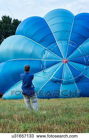 Stock Image - Todi, Italy: July 19, 2013; Todi Mongolfiere Hot Air Balloon Festival - red and blue hot air balloon with star pattern being inflated, with woman holding cord as it rises in Middle Tiber Valley field, in early morning sunshine, close up view, Umbria, It. Fotosearch