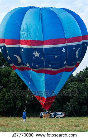 Stock Image - Todi, Italy: July 19, 2013; Todi Mongolfiere Hot Air Balloon Festival - red and blue hot air balloon with star pattern being inflated, with woman holding cord as it rises in Middle Tiber Valley field, in early morning sunshine, Umbria, Italy. Fotosearch