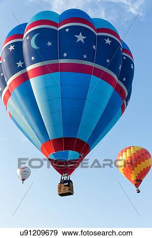 Stock Photo - Todi, Italy: July 9, 2013 - Todi Mongolfiere Hot Air Balloon Festival; red and blue hot air balloon in sky with colourful balloons on either side beyond in blue sky above, Umbria, Italy. Fotosearch