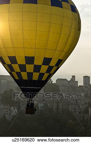 Stock Image - Todi Mongolfiere Hot Air Balloon Festival - blue and yellow checkered hot air balloon over medieval hilltop town of Todi in early morning hazy sunshine, aerial view, Umbria, Italy. Fotosearch