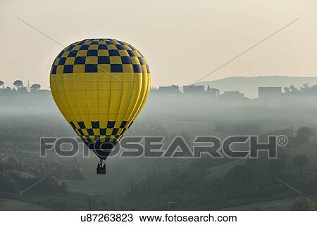 Todi Mongolfiere Hot Air Balloon Festival - blue and yellow checkered hot air balloon over Middle Tiber Valley with hills layered in early morning hazy sunshine, aerial view, Umbria, Italy View Large Photo Image Stock Image - Todi Mongolfiere Hot Air Balloon Festival - blue and yellow checkered hot air balloon over Middle Tiber Valley with hills layered in early morning hazy sunshine, aerial view, Umbria, Italy. Fotosearch