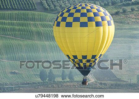 Stock Photo - Todi Mongolfiere Hot Air Balloon Festival; blue and yellow checkered hot air balloon in sky over green undulating fields in Middle Tiber Valley in early morning sunshine, aerial view, Umbria, Italy. Fotosearch