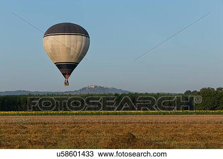 Stock Image - View of black and white hot air balloon over freshly mown hay fields, sunflowers beyond, and hilltop town of Todi in distance, sunny early morning, Middle Tiber Valley, Umbria, Italy. Fotosearch