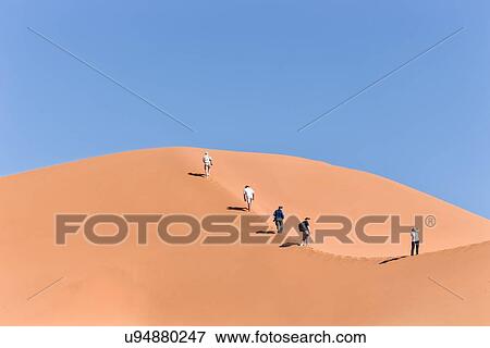Stock Photo - View of five people walking up line of red sand dune with different tones in shadow of early morning sunlight, and sand being blown by winds, blue sky above, Namib desert, Namibia, South West Africa. Fotosearch