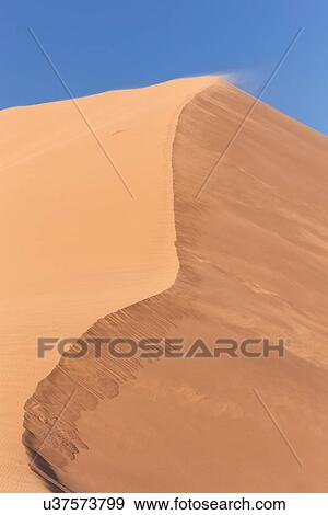 View of line of red sand dune with different tones in shadow of early morning sunlight, and sand being blown by winds, blue sky above, Namib desert, Namibia, South West Africa View Large Photo Image Stock Photo - View of line of red sand dune with different tones in shadow of early morning sunlight, and sand being blown by winds, blue sky above, Namib desert, Namibia, South West Africa. Fotosearch