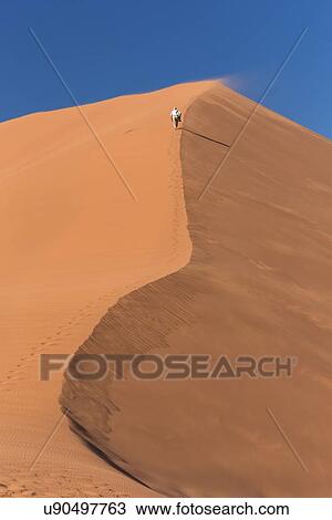 View of man running up line of red sand dune with different tones in shadow of early morning sunlight, and sand being blown by winds, blue sky above, Namib desert, Namibia, South West Africa View Large Photo Image Stock Image - View of man running up line of red sand dune with different tones in shadow of early morning sunlight, and sand being blown by winds, blue sky above, Namib desert, Namibia, South West Africa. Fotosearch