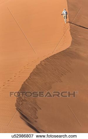 View of man running up line of red sand dune with different tones in shadow of early morning sunlight, and sand being blown by winds, Namib desert, Namibia, South West Africa View Large Photo Image Stock Photo - View of man running up line of red sand dune with different tones in shadow of early morning sunlight, and sand being blown by winds, Namib desert, Namibia, South West Africa. Fotosearch