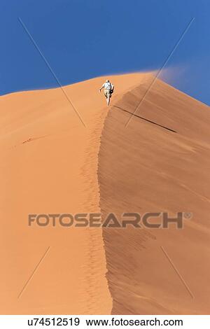 Stock Photo - View of man running up line of red sand dune with different tones in shadow of early morning sunlight, and sand being blown by winds, blue sky above, Namib desert, Namibia, South West Africa. Fotosearch