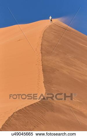View of man running up line of red sand dune with different tones in shadow of early morning sunlight, and sand being blown by winds, blue sky above, Namib desert, Namibia, South West Africa View Large Photo Image Stock Photography - View of man running up line of red sand dune with different tones in shadow of early morning sunlight, and sand being blown by winds, blue sky above, Namib desert, Namibia, South West Africa. Fotosearch