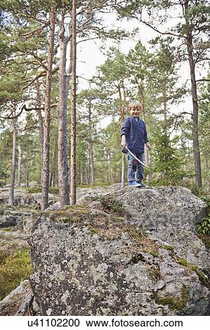 Stock Image - Boy standing on rocks holding stick. Fotosearch