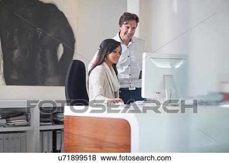 Businesswoman sitting at desk using computer, man behind her View Large Photo Image Stock Photo - Businesswoman sitting at desk using computer, man behind her. Fotosearch