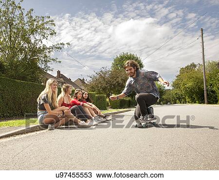 Stock Image - Four young women sitting on kerb watching skateboarder. Fotosearch