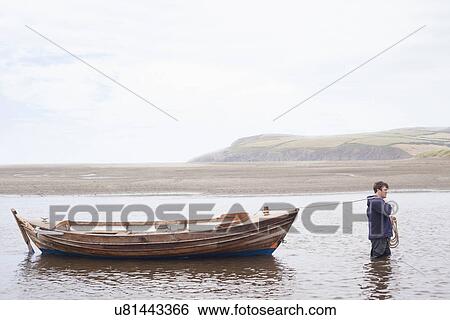 Stock Photograph - Man pulling rowing boat through water, Wales, UK. Fotosearch
