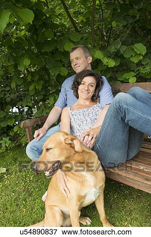 Stock Photo - Mature couple sitting on garden bench with dog. Fotosearch