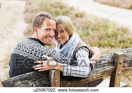 Stock Image - Mid adult couple on wooden bench looking at camera. Fotosearch