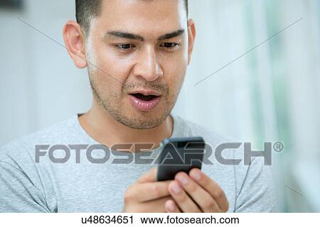 Stock Image - Mid adult man looking surprised at smartphone. Fotosearch