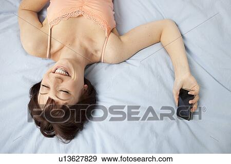 Stock Photo - Overhead view of young woman lying on bed with mobile phone. Fotosearch