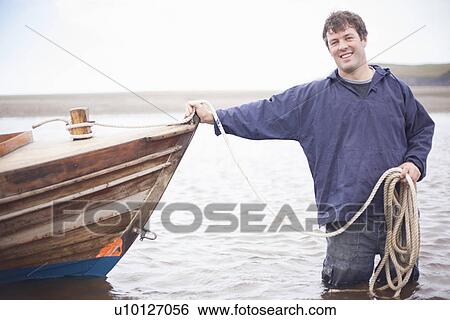 Stock Photograph - Portrait of man holding rope leaning on rowing boat, Wales, UK. Fotosearch