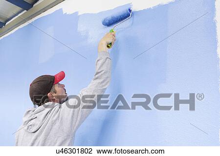Stock Image - Rear view of man painting wall blue. Fotosearch