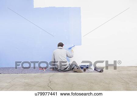 Stock Image - Rear view of man painting wall blue. Fotosearch