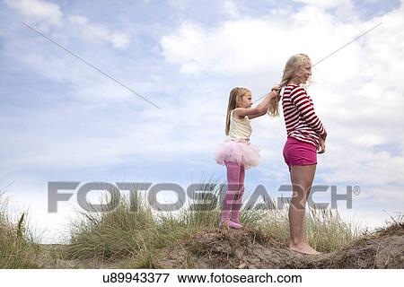 Stock Photo - Two girls on dunes, one plaiting friend's hair, Wales, UK. Fotosearch