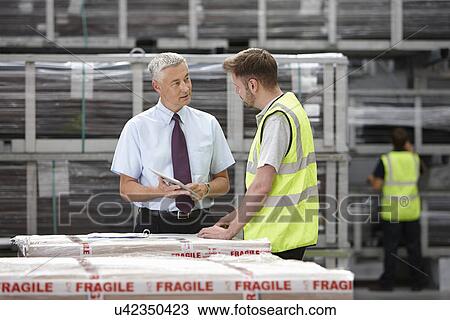 Stock Image - Warehouse worker and manager discussing order in engineering warehouse. Fotosearch