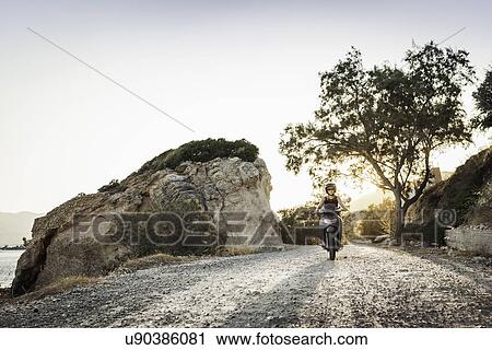 Stock Image - Woman riding moped along coast at Pythagorio, Samos, Greece. Fotosearch