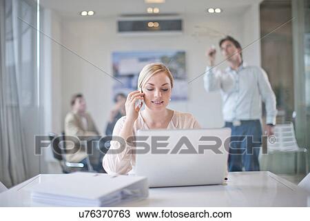 Woman sitting at conference room table using laptop and mobile phone, colleagues working in background View Large Photo Image Stock Image - Woman sitting at conference room table using laptop and mobile phone, colleagues working in background. Fotosearch