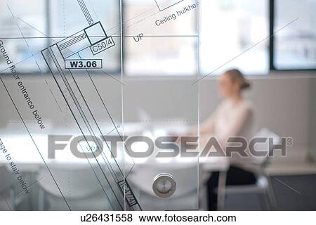 Stock Photo - Woman sitting at conference room table using laptop, view through window and architect plans. Fotosearch