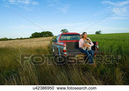 Stock Image - Woman sitting on back of pick up truck in a field, holding a dog. Fotosearch