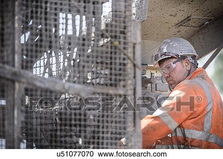 Stock Image - Worker in hard hat operating drilling rig in field. Fotosearch