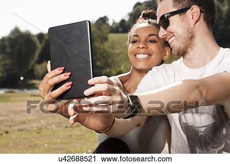 Stock Image - Young couple using digital tablet to photograph themselves. Fotosearch