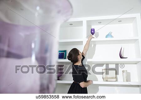 Stock Photo - Young woman reaching for wine glass on shelf in living room. Fotosearch