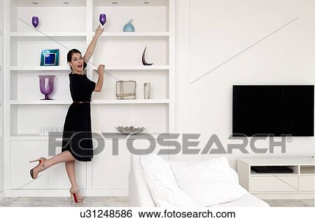 Stock Photograph - Young woman reaching for wine glass on shelf in living room. Fotosearch