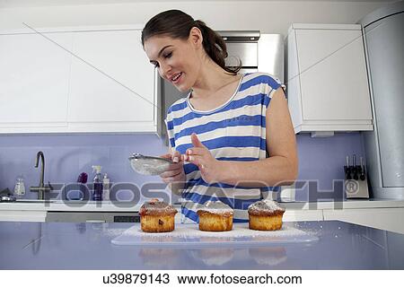 Stock Image - Young woman sieving icing sugar over cakes. Fotosearch