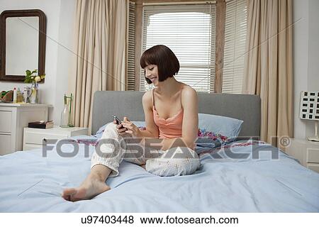 Young woman sitting on bed looking at mobile phone View Large Photo Image Stock Photo - Young woman sitting on bed looking at mobile phone. Fotosearch