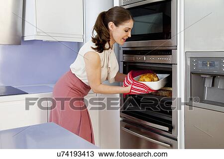 Stock Image - Young woman taking roasting dish out of oven. Fotosearch