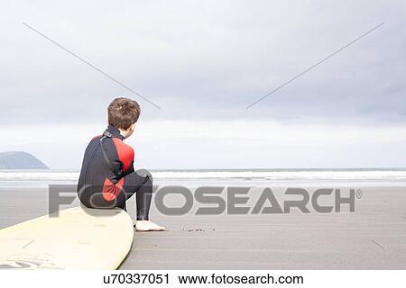 Stock Image - Boy sitting on surfboard on beach. Fotosearch