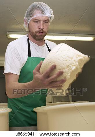 Stock Image - Cheesemaker holding part processed cheese round at farm factory. Fotosearch