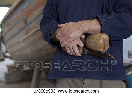 Stock Image - Close up of carpenters hands holding chisel in boat workshop. Fotosearch