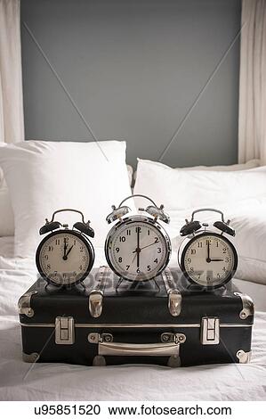 Stock Image - Closed suitcase on bed with three alarm clocks. Fotosearch