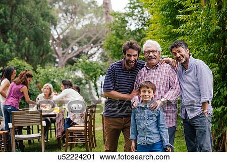 Stock Image - Family at gathering in garden - male family members in foreground, female family members at table in background. Fotosearch