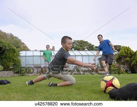 Stock Image - Father and sons playing football in garden. Fotosearch