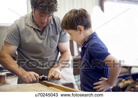 Stock Image - Father showing son carpentry in boat workshop. Fotosearch