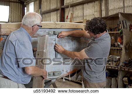 Stock Image - Father watching son restore boat in workshop. Fotosearch