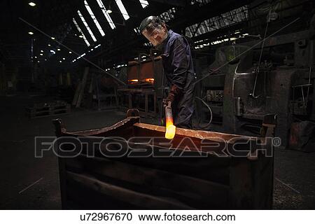Stock Image - Forge worker preparing to release finished upset forging into steel container. Fotosearch