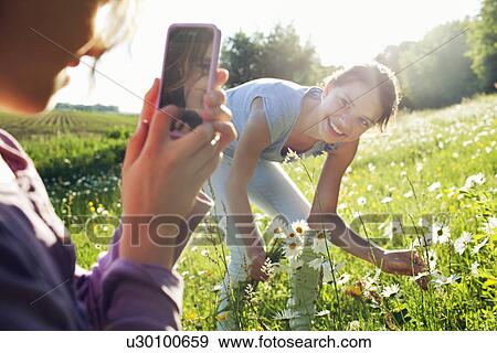 Stock Photo - Girl taking photograph of girl picking flowers. Fotosearch
