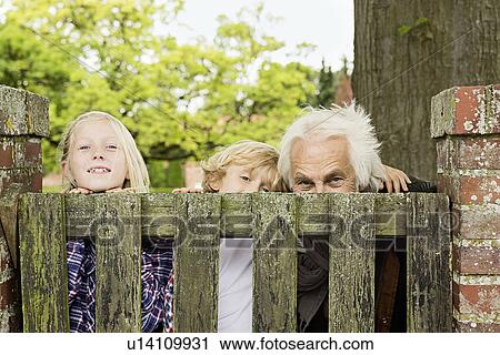 Stock Image - Grandfather and grandchildren peering over wooden gate. Fotosearch