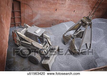 Stock Image - High angle view of digger and grab unloading metal alloy in ship's hull. Fotosearch